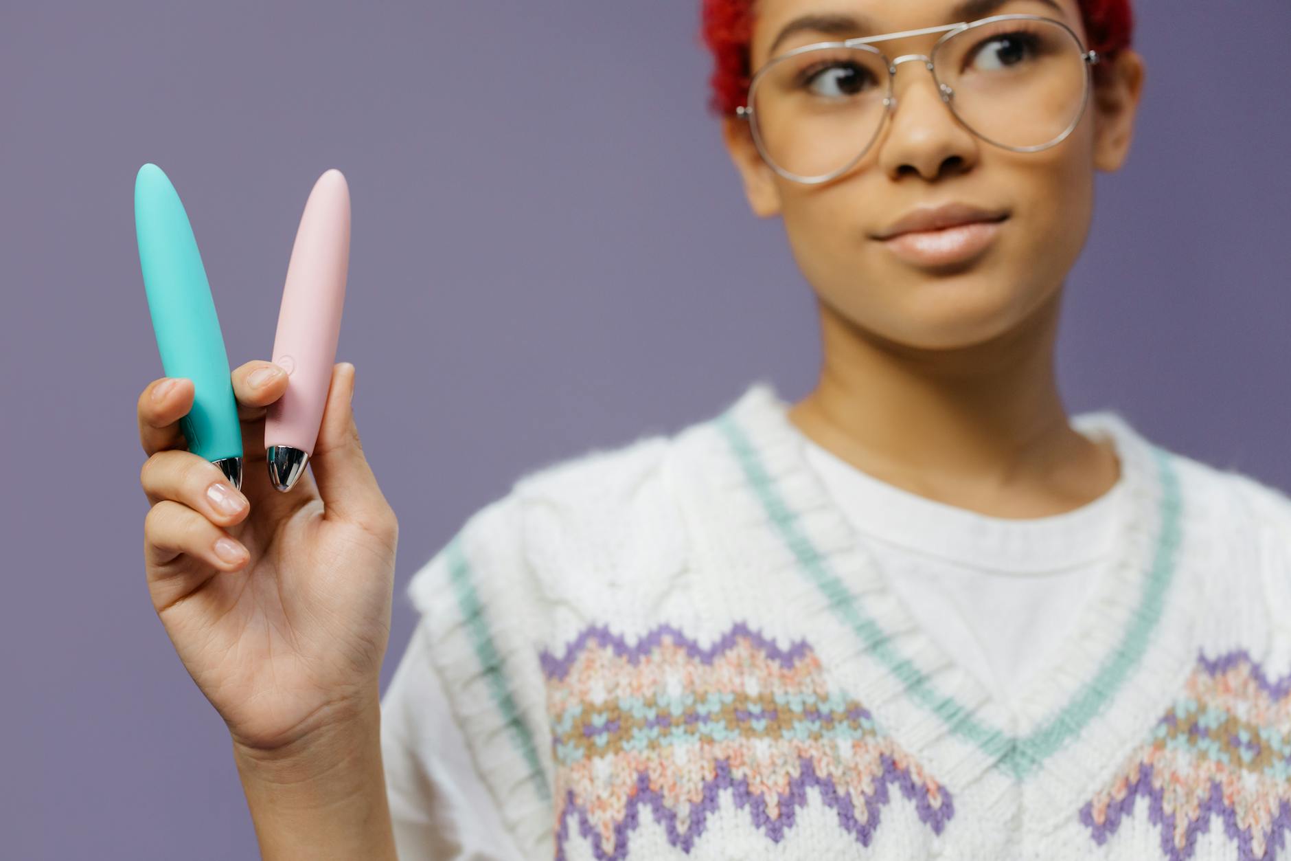 Eine junge Frau mit Brille hält zwei bunte Vibratoren vor lila Hintergrund