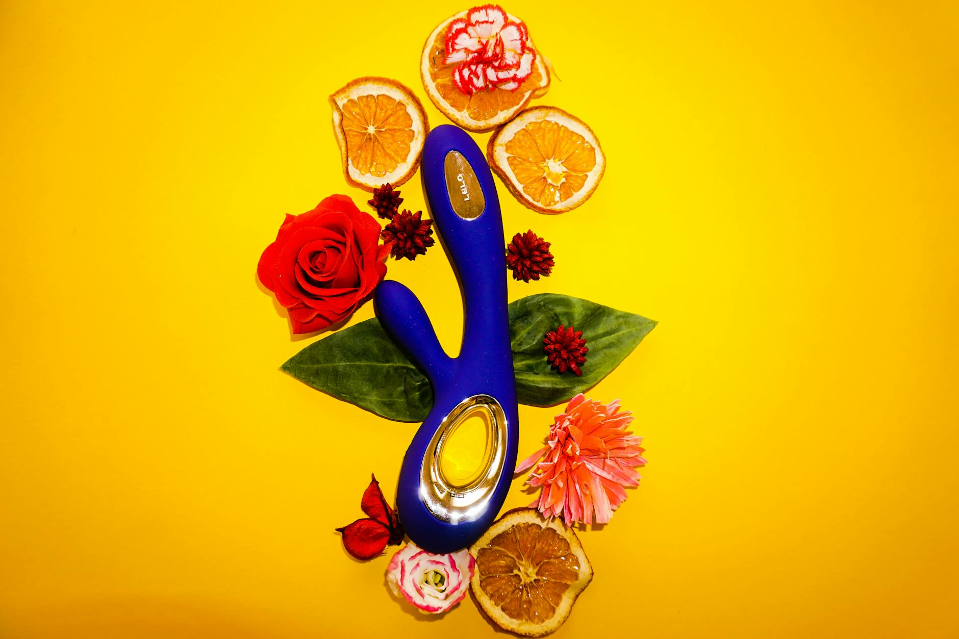 A vibrant blue lemon clitoral vibrator surrounded by fresh flowers and citrus slices on a bright yellow background