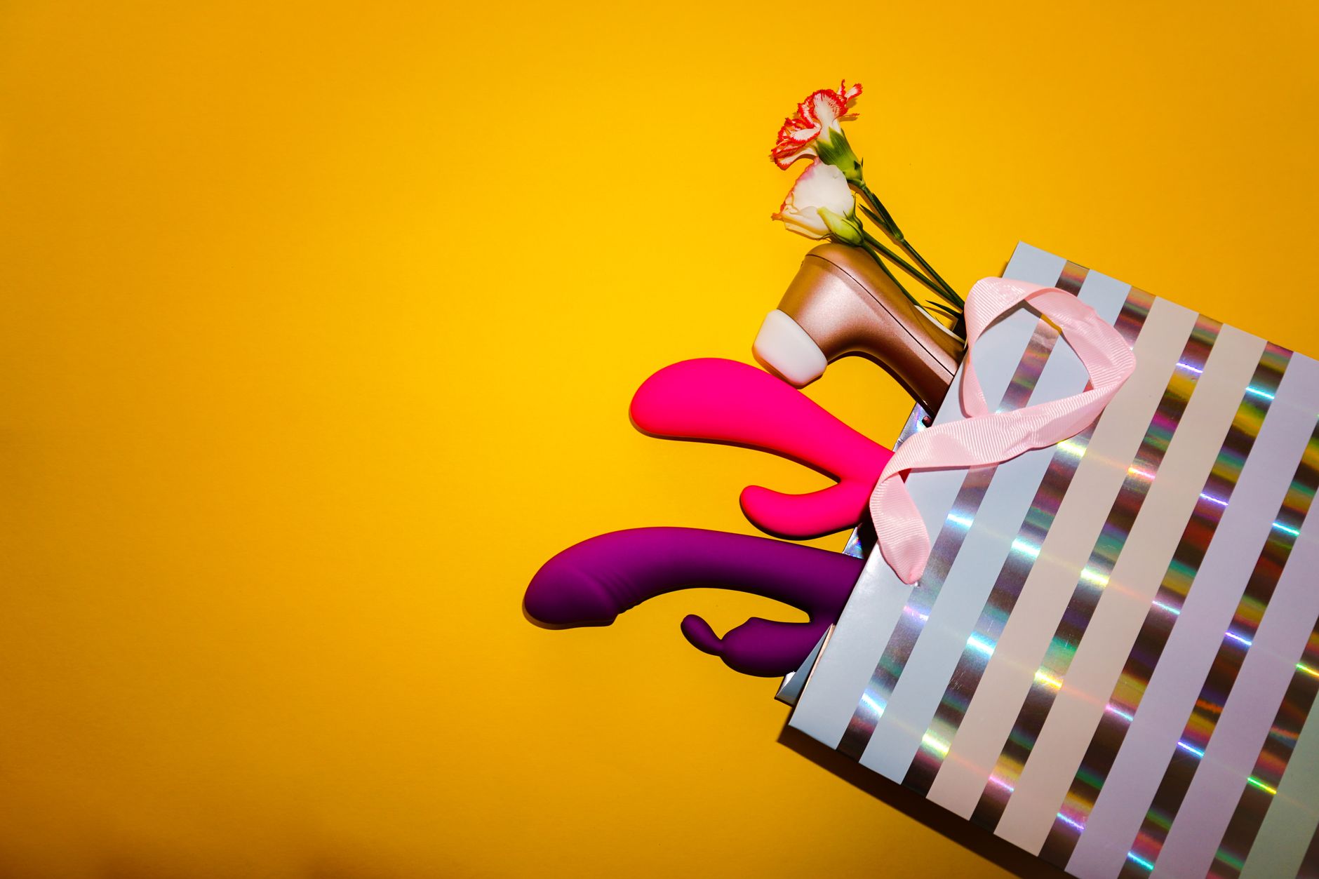 Farbenfrohe Vibratoren mit Blüten in einer holografischen Geschenktasche vor leuchtend gelbem Hintergrund.